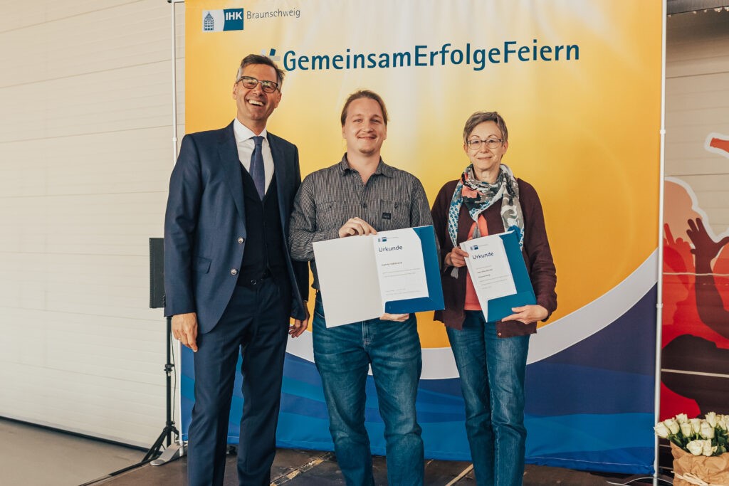 Ein junger Mann steht zwischen dem IHK-Präsidenten und seiner Ausbilderin und zeigt fröhlich seine Auszeichnung