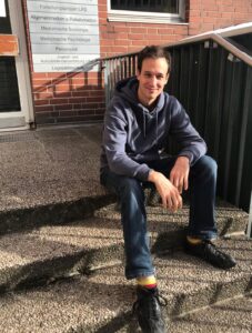 Ein junger Mann stitz vor einer Eingangstür auf der obersten Treppenstufe und lächelt